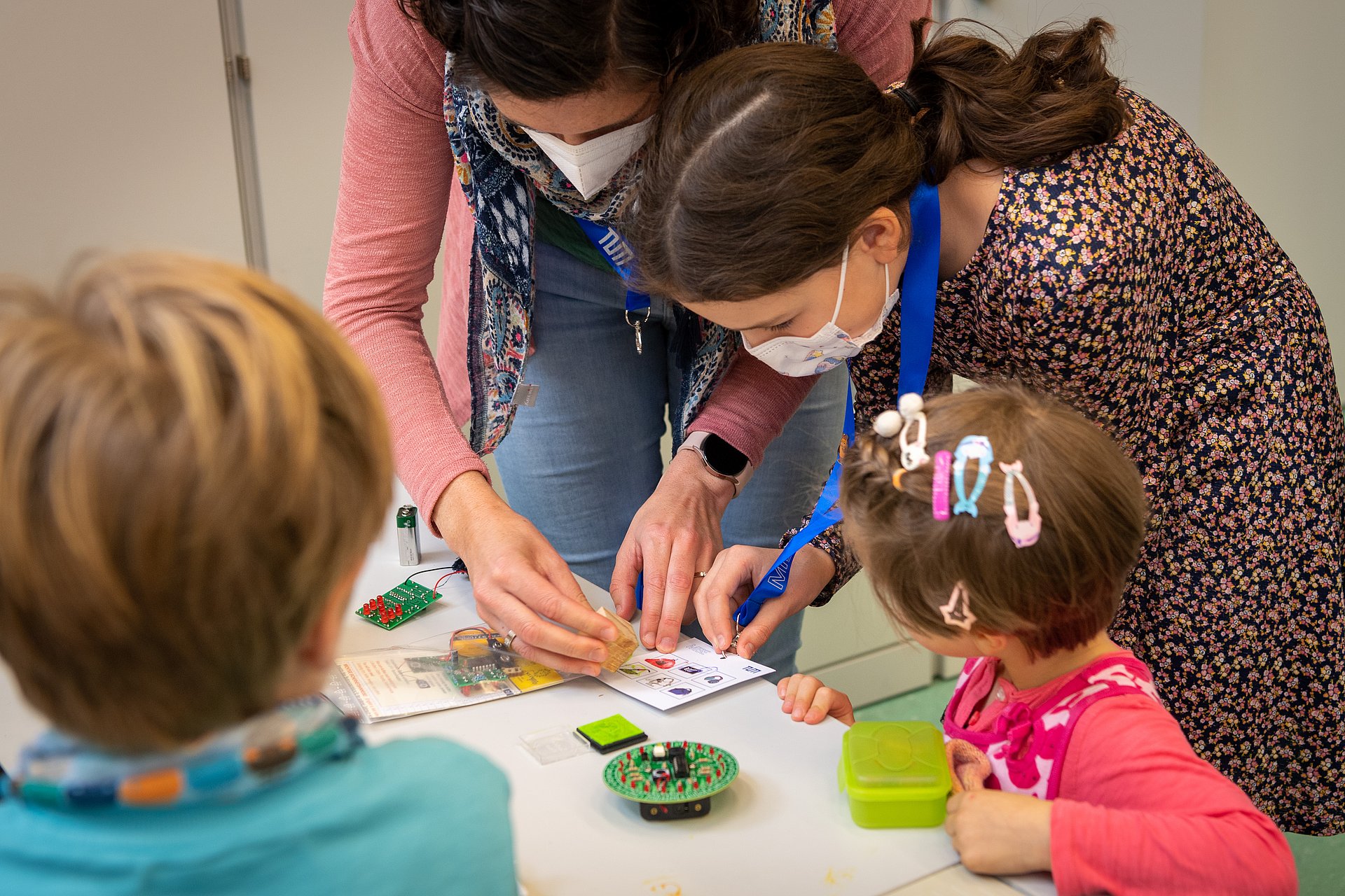 Adults with masks and children at a table. A woman stamps a card at a hands-on station.