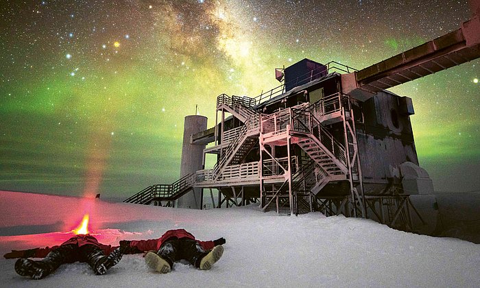Astroteilchenphysiker Martin Wolf und sein Kollege liegen auf dem Rücken vor dem  IceCube-Teleskop im Schnee und blicken in den Himmel, wo man die Milchstraße klar sieht. 