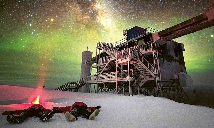 Astroparticle physicist Martin Wolf and his colleague lie on their backs in the snow in front of the IceCube telescope and look up at the sky, where the Milky Way can be seen clearly. 