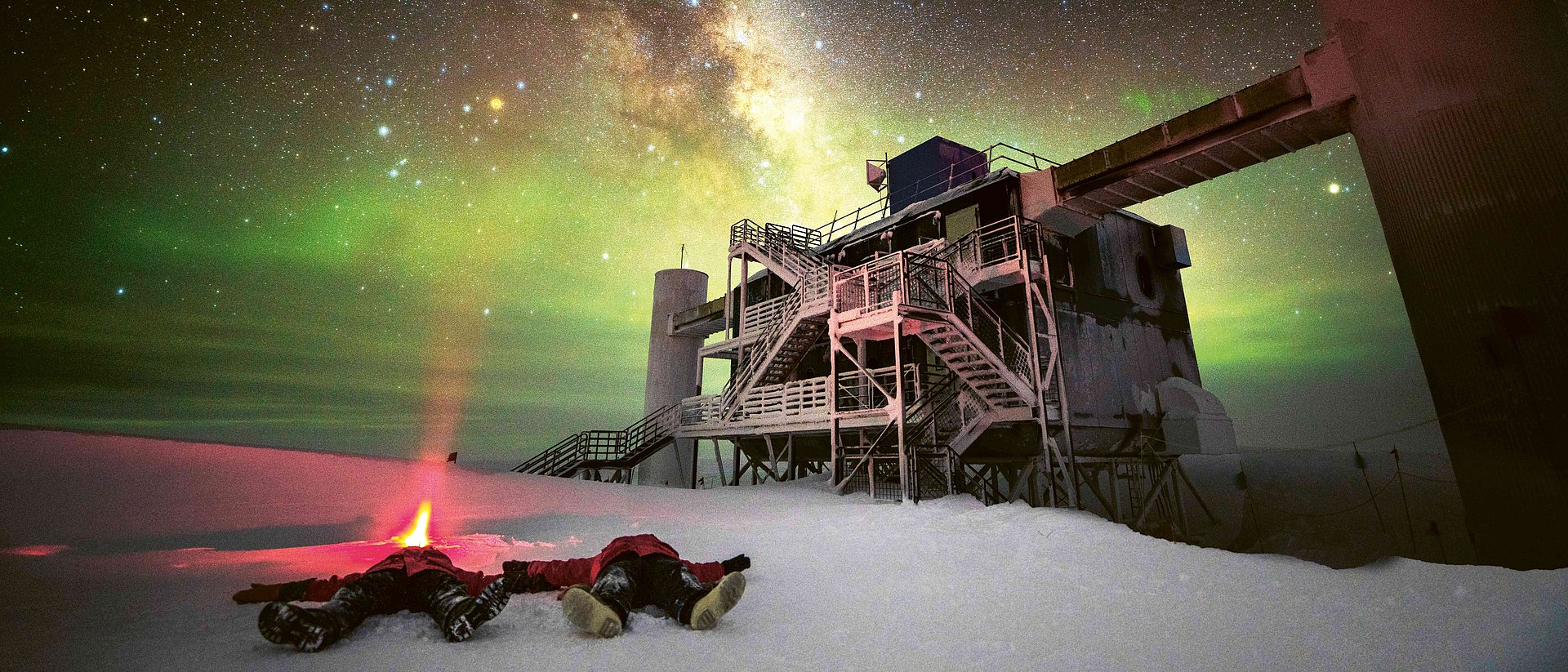 Astroteilchenphysiker Martin Wolf und sein Kollege liegen auf dem Rücken vor dem  IceCube-Teleskop im Schnee und blicken in den Himmel, wo man die Milchstraße klar sieht. 