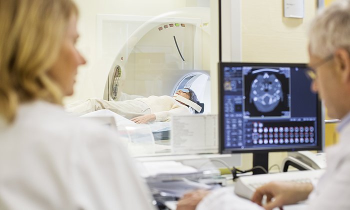 Two people are sitting in front of a screen. A third person lies in the background on a stretcher of a magnetic resonance tomograph.