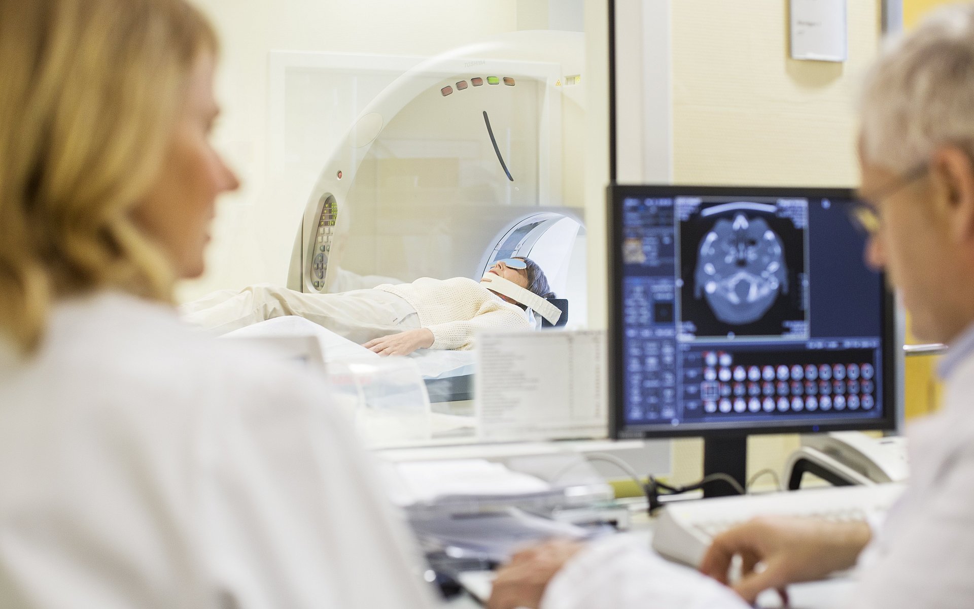 Two people are sitting in front of a screen. A third person lies in the background on a stretcher of a magnetic resonance tomograph.