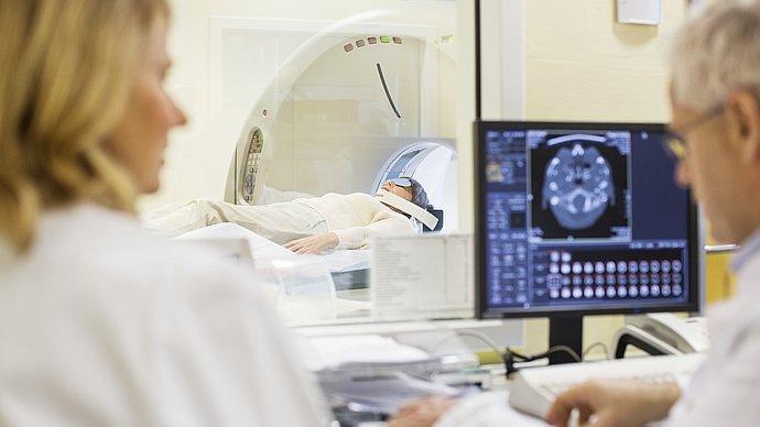 Zwei Personen sitzen vor einem Bildschirm. Eine dritte Person liegt im Hintergrund auf einer Bahre eines Magnetresonanztomographen.
