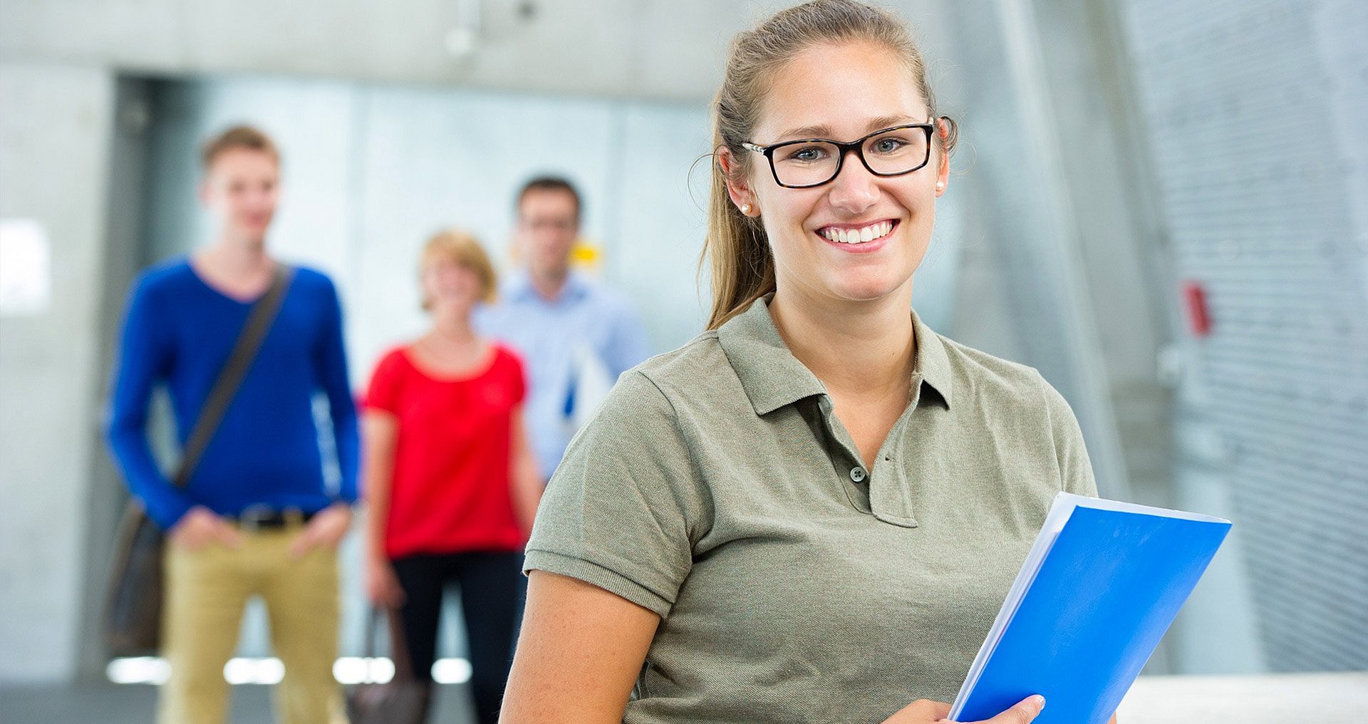 A female student is standing in the room, smiling and holding a blue folder in her hand, while three other students are visible out of focus in the background.