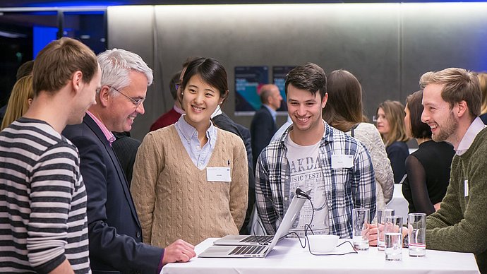 Teilnehmende des IDEAward tauschen sich aus. Fünf Menschen, die an einem Tisch zusammenstehen und gemeinsam lachen. Auf dem Tisch stehen zwei Laptops und mehrere Wasserglässer.