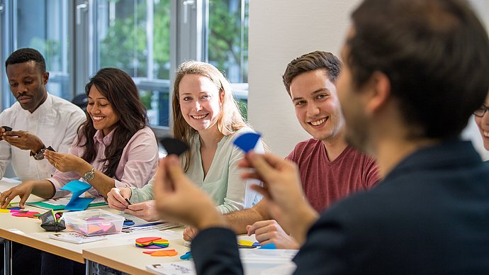 Four students in a seminar room, in the foreground a lecturer out of focus.