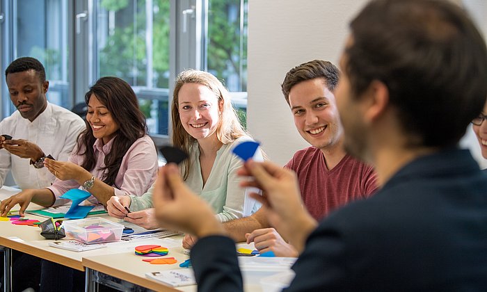 Four students in a seminar room, in the foreground a lecturer out of focus.