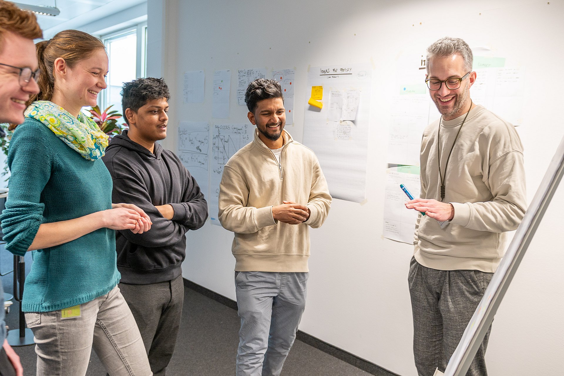 Vier Studierende blicken auf einen Aufsteller. Der Leiter des Teams steht neben dem Austeller und erklärt, was zu sehen ist.