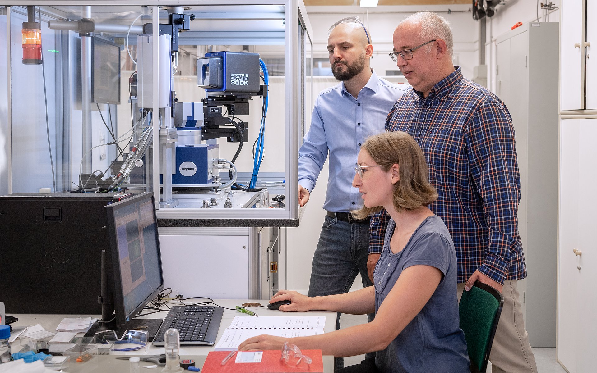 Das Foto zeigt Dr. Venturini (links), Prof. Nilges und Erstautorin Anna Vogel vor einem Messgerät zur Bestimmung von Kristallstrukturen, einem sogenannten Einkristalldiffraktometer.