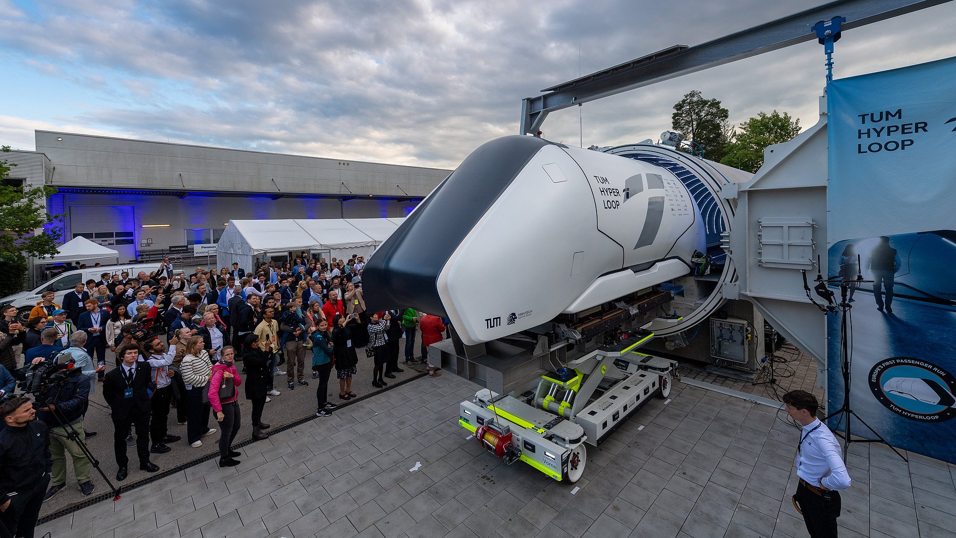 A Hyperloop Pod is on a transport rack in front of a concrete tube. Many guests are standing next to it.