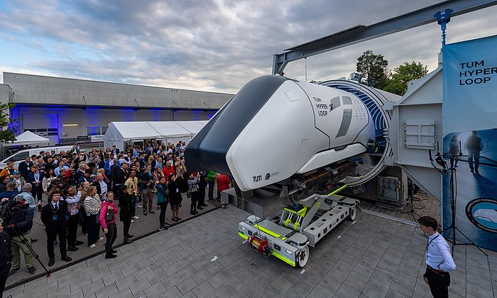 Ein Hyperloop Pod ist auf einem Transportgestell vor einer Betonröhre. Viele Gäste stehen daneben.