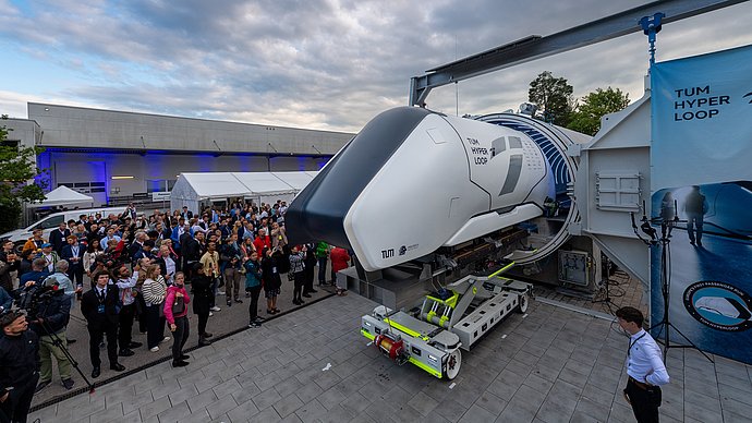 Ein Hyperloop Pod ist auf einem Transportgestell vor einer Betonröhre. Viele Gäste stehen daneben.