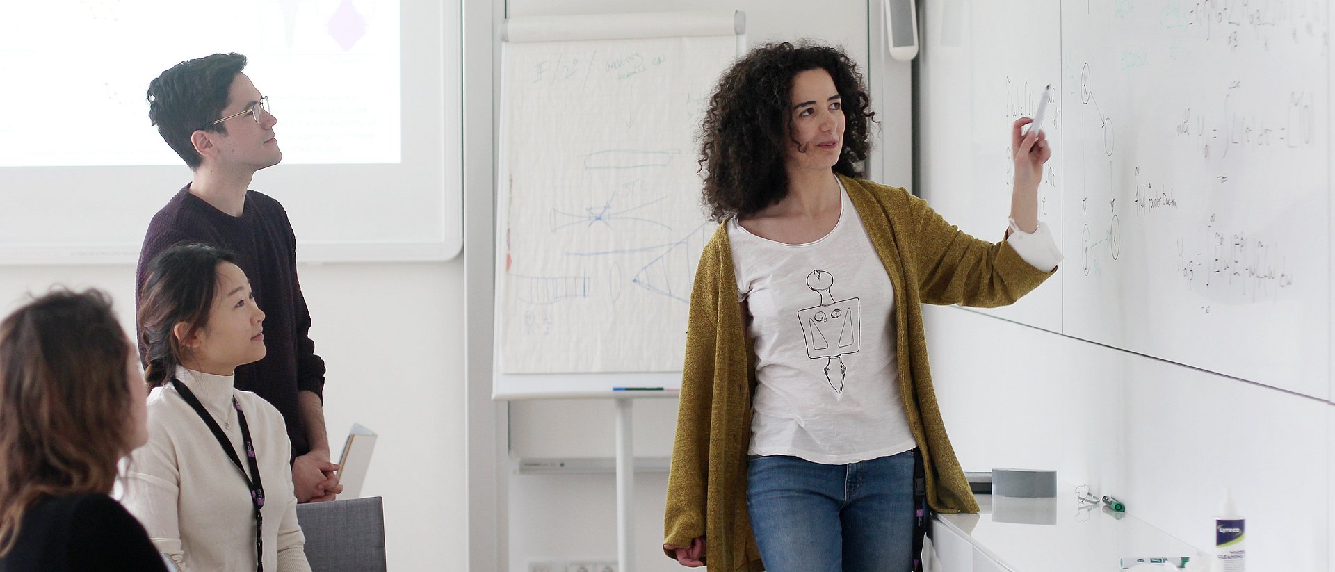  In a seminar room, a female scientist explains a formula written on a whiteboard to three young colleagues.