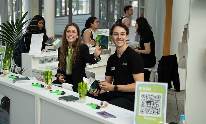 eine Studentin und ein Student lächeln hinter dem Infotresen der Messe IKOM entgegen. 