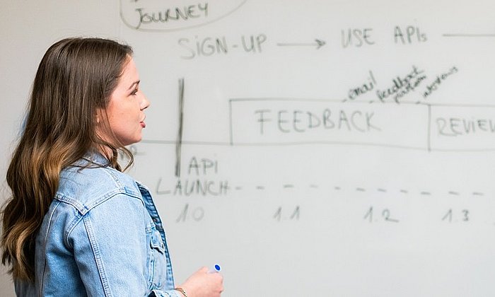 Young woman stands in front of a whiteboard with various processes outlined in handwriting and the words "feedback" and "review"