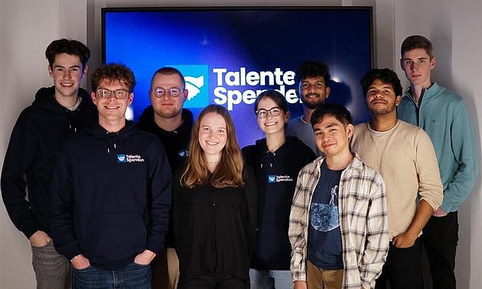 neun Mitglieder der Initiative Talende Spenden stehen als Gruppe zusammen und lächeln freundlich in die Kamera. die Männer und Frauen sthene vor einem großen Monitor, auf dem das Logo und der Schriftzug Talente Spenden zu sehen sind.