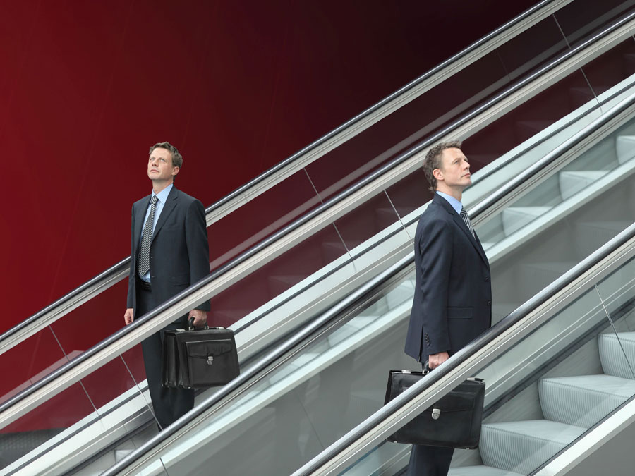 Je ähnlicher sich Vorstandschef und Vorgänger sind, desto eher schlägt der Neue einen anderen Weg ein. (Bild: iStockphoto.com / Visivasnc) Männer in Anzügen auf Rolltreppen in entgegengesetzten Richtungen