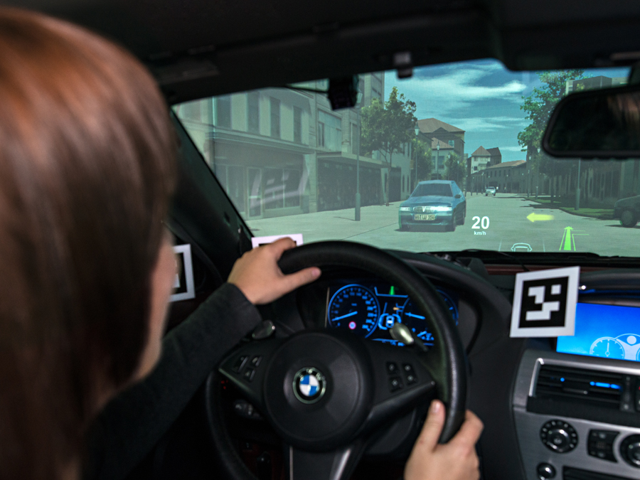 Eine Frau sitzt im Fahrzeugsimulator und blickt auf die Leinwand, auf der das Head-up-Display zu sehen ist.