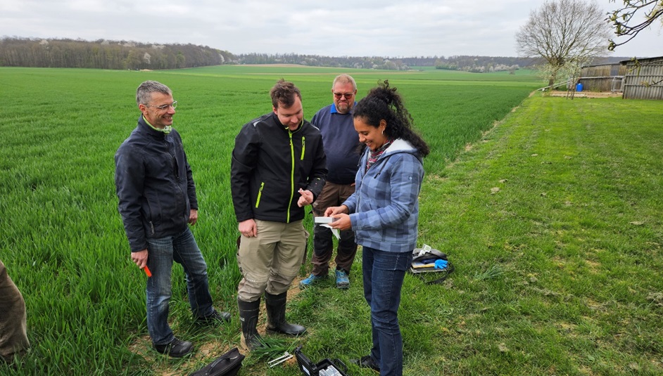 Start-up NutriSen bringt Labor auf den Acker Eine Frau und drei Männer stehen auf einem Acker. Die Frau zeigt den Männern ein Gerät in ihrer Hand. Es handelt sich um das Gerät, dass das NutriSen-Team zur smarten Düngung entwickelt hat.