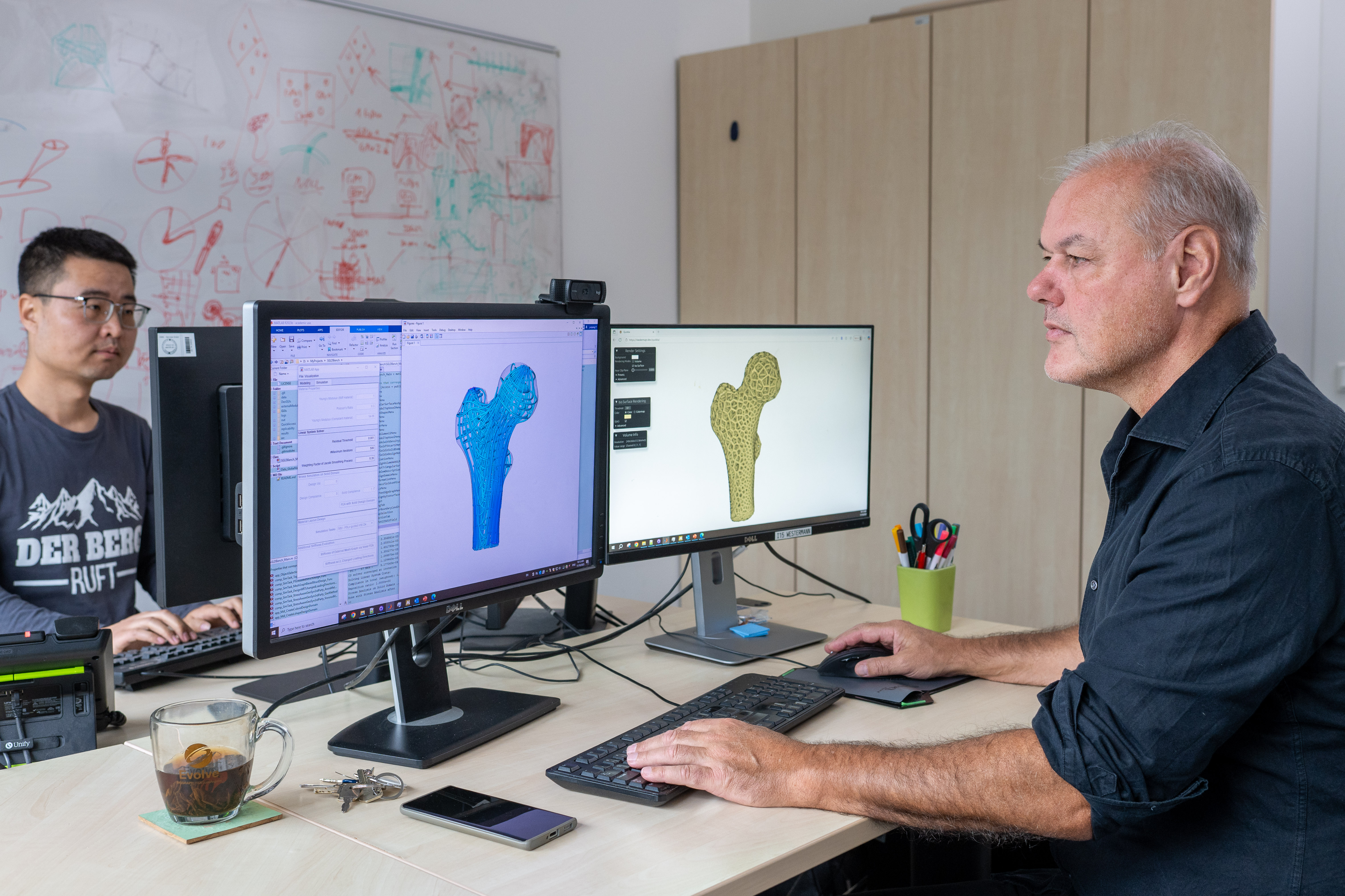 Zwei Forschende vor ihren Computern werden dargestellt. Auf den Bildschirmen sind zwei Hüftimplantate abgebildet, die mithilfe unterschiedlicher Ansätze konstruiert wurden.
