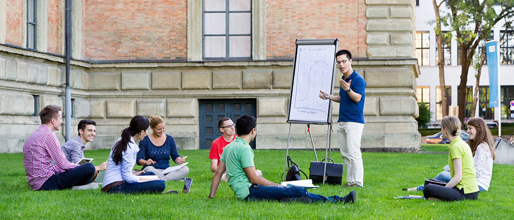 Studierende sitzen am Campus auf einer Wiese und diskutieren ein Konzept, das auf einem Whiteboard skizziert wird.